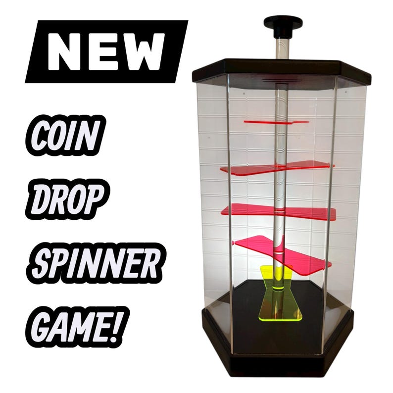 Puede incluir: Un juego de monedas giratorias de forma hexagonal transparente con componentes internos rojos y amarillos. El juego tiene una base y una parte superior negras, y el texto "NEW COIN DROP SPINNER GAME!" se muestra en blanco y negro.