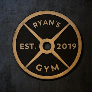 Op de afbeelding: Een zwart en gouden houten gewichtsplaat bord met de tekst "Ryan's Gym Est. 2019"
