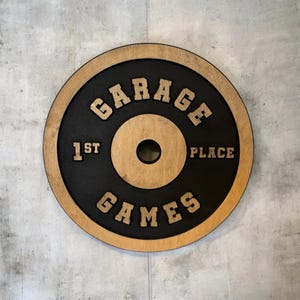 Op de afbeelding: Een zwarte en bruine houten gewichtsplaat met de tekst "Garage Games 1st Place" in gouden letters.