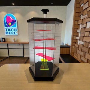 Puede incluir: Una vitrina hexagonal transparente con base y tapa negras. En el interior, hay varios estantes rojos y una estructura de soporte amarilla. La vitrina est&aacute; sobre una mesa de madera de color claro, con un letrero de Taco Bell al fondo.