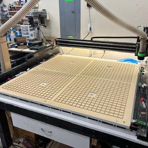 May include: A large CNC router machine featuring a light tan MDF spoilboard with a grid pattern of square holes. The machine has a black metal frame, a white drawer base, and a flexible dust collection hose attached to the cutting head assembly.