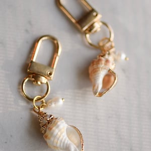 Pode incluir: Dois chaveiros dourados com um charme de concha e uma pérola. As conchas são brancas com listras marrons.