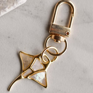 Puede incluir: Llavero dorado con un dije de manta raya de nácar blanco e iridiscente.