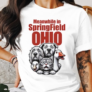 Puede incluir: Camiseta blanca con el texto "Meanwhile in Springfield OHIO" en letras rojas. La camiseta presenta una ilustración de dibujos animados de un perro, un gato, un ganso y otro perro.