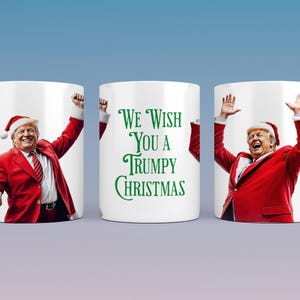 Könnte beinhalten: Weiße Keramiktasse mit festlichem Design. Die Tasse zeigt eine Person in einem Weihnachtsmannkostüm und -hut mit dem Text "We Wish You a Trumpy Christmas" in Grün. Die Person lächelt und hebt die Arme.