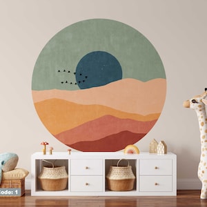 May include: A round wall decal featuring a stylised landscape design with a blue circle, orange, yellow and red hills, and a flock of black birds flying across the sky. The decal is on a white wall above a white dresser with two drawers.