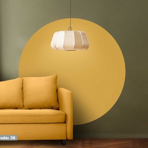 Puede incluir: Un sofá amarillo con tres cojines se encuentra frente a una pared pintada en dos tonos de verde. Un círculo amarillo redondo está pintado en la pared. Una lámpara colgante blanca cuelga del techo sobre el sofá.