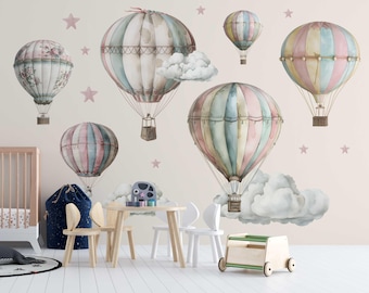 May include: A nursery wall decal set featuring watercolor-style hot air balloons in shades of pink, blue, and beige. The balloons are decorated with stripes and patterns, and some have clouds around them. There are also pink stars scattered throughout the design.