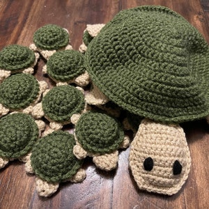 May include: A collection of crocheted green and beige turtle toys. The turtles are arranged in a circle with a larger turtle in the center. The turtles have detailed features, including eyes, legs, and shells.