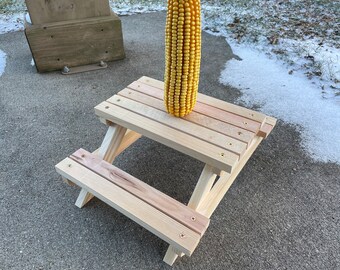 May include: A small wooden picnic table with a yellow corn cob on top. The table is made of unfinished wood and has a simple design.