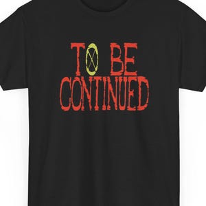May include: Black t-shirt with the phrase "TO BE CONTINUED" in red, distressed lettering. The word "TO" and "CONTINUED" are stacked, with a yellow and green symbol between them. The shirt is a classic crew neck style.