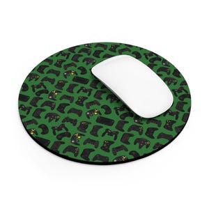 May include: A round green mousepad featuring a pattern of black video game controllers. A white computer mouse rests on the mousepad. The controllers have yellow and green accents. The mousepad is designed for computer use.