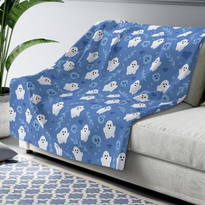 May include: A blue throw blanket with a pattern of white ghosts, skulls, hearts, and leaves. The blanket is draped over a white couch. The blanket is made of a soft material.