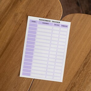 K&ouml;nnte beinhalten: Ein wei&szlig;er Aufgaben-Tracker mit lavendelfarbenem Rand und &Uuml;berschriften. Der Text "Assignment Tracker" steht oben, mit Spalten f&uuml;r Kurs, Aufgabe, F&auml;lligkeitsdatum und Fertigstellung. Der Tracker liegt auf einer Holzoberfl&auml;che.