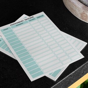 以下が含まれることがあります： 「Assignment Tracker」の文字が上部に印刷された、水色のグリッドデザインの白い宿題トラッカーシートが2枚。シートは黒い表面に重ねられています。