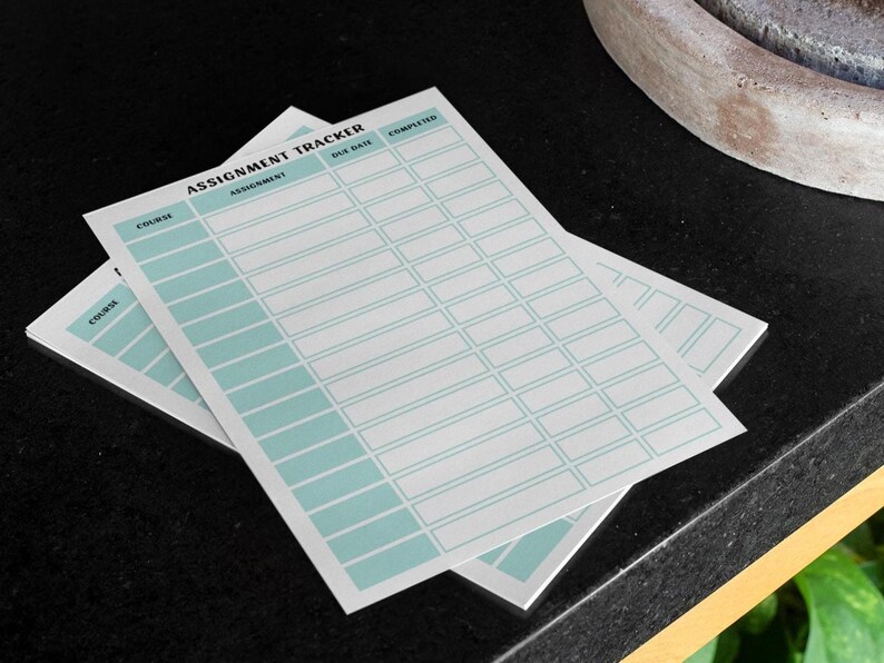 以下が含まれることがあります： 「Assignment Tracker」の文字が上部に印刷された、水色のグリッドデザインの白い宿題トラッカーシートが2枚。シートは黒い表面に重ねられています。
