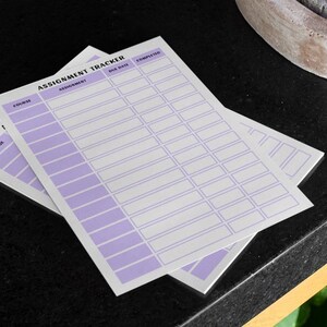 K&ouml;nnte beinhalten: Zwei wei&szlig;e Aufgabenverfolgungsbl&auml;tter mit einem lila Design. Die Bl&auml;tter tragen oben die Worte "Assignment Tracker" mit Spalten f&uuml;r "Kurs", "Aufgabe", "F&auml;lligkeitsdatum" und "Abgeschlossen". Die Bl&auml;tter sind auf einer schwarzen Oberfl&auml;che gestapelt.