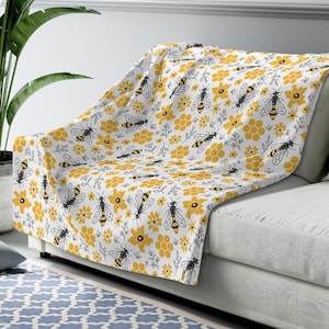 May include: A white throw blanket with a repeating pattern of yellow flowers, honeycombs, and bees. The bees are black and yellow, and the flowers have a simple, stylized design. The blanket is draped over a gray couch.