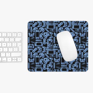 Puede incluir: Alfombrilla de ratón negra con un patrón de mandos y consolas de videojuegos azules. Un ratón de ordenador blanco está sobre la alfombrilla, junto a un teclado blanco.