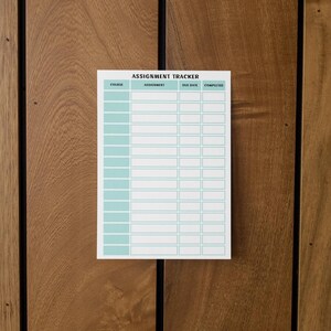 以下が含まれることがあります： 上部に「ASSIGNMENT TRACKER」というタイトルが付いた、ターコイズ色のグリッドの白い課題トラッカー。トラッカーは木製の表面にあります。「COURSE」、「ASSIGNMENT」、「DUE DATE」、「COMPLETED」とラベル付けされた列があります。