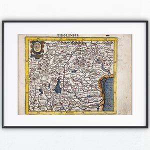 Op de afbeelding: Ingelijste vintage kaartprint van Tirol, met het woord "TIROLENSIS" bovenaan. De kaart bevat gedetailleerde illustraties van bergen, rivieren en steden, met een gele rand en een decoratief embleem. De print zit in een zwart frame.