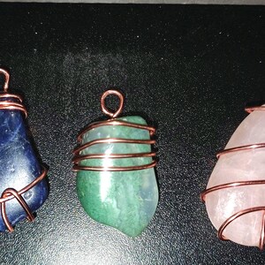 May include: Three gemstone pendants. One is a deep blue stone, one is green, and the last is a pale pink. Each is wrapped with copper wire, forming a loop at the top for hanging. The pendants are on a dark surface.
