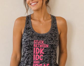 May include: A charcoal gray racerback tank top with a distressed, mottled texture. The front features bold, bright pink text that reads: STUCK BETWEEN IDK IDC AND IDGAF, arranged in a vertical block format with a rounded neckline and sleeveless cut.