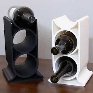 Puede incluir: Dos porta botellas de vino, uno negro y otro blanco, cada uno con tres ranuras para botellas. El porta botellas negro tiene una botella en la ranura superior, mientras que el porta botellas blanco tiene dos botellas en las dos ranuras inferiores.