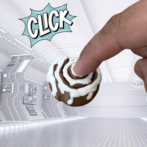 May include: A brown cinnamon roll with white icing is held by a finger. A comic book style speech bubble with the word "CLICK" in white letters with a blue outline is in the background. The background is a futuristic white room.