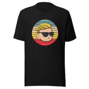 May include: Black t-shirt featuring a retro-style graphic of a cartoon face wearing sunglasses, set against a circular background with horizontal stripes in yellow, red, blue, and white. The design has a distressed, vintage look.