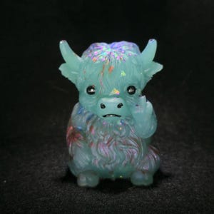 May include: A small, translucent light blue figurine of a highland cow with iridescent glitter flakes embedded throughout its body. The figure features textured fur, two small horns, dark eyes, and a snout, with one hoof raised near its face.
