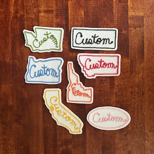 May include: Seven felt patches in various shapes and colors, each with the word "Custom" embroidered in a different color thread. The patches are shaped like the states of Washington, Oregon, Idaho, Montana, California, and a circle.
