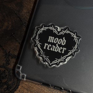 Könnte beinhalten: Ein schwarzer, herzförmiger Handyhalter mit den Worten "mood reader" in weißer Schrift. Das Herz ist von einem Stacheldrahtmuster umgeben. Der Halter befindet sich auf einer transparenten Handyhülle.