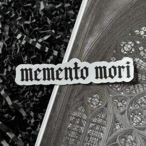 K&ouml;nnte beinhalten: Ein wei&szlig;er Aufkleber mit dem Schriftzug "memento mori" in gotischer Schrift. Der Aufkleber ist diagonal &uuml;ber einem Schwarz-Wei&szlig;-Bild eines Kathedralenfensters und schwarzem geschreddertem Papier platziert.
