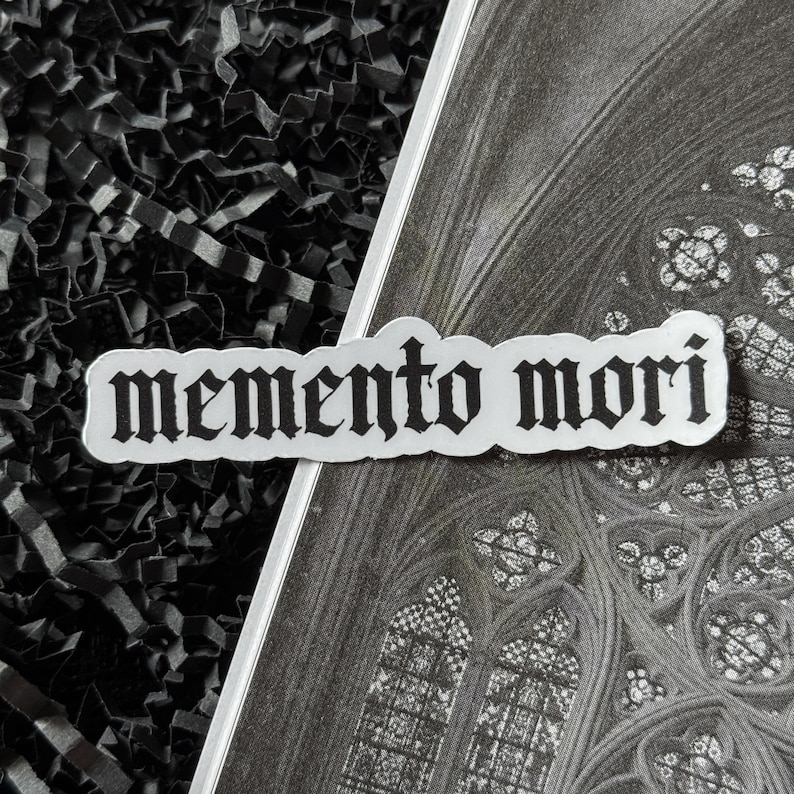 K&ouml;nnte beinhalten: Ein wei&szlig;er Aufkleber mit dem Schriftzug "memento mori" in gotischer Schrift. Der Aufkleber ist diagonal &uuml;ber einem Schwarz-Wei&szlig;-Bild eines Kathedralenfensters und schwarzem geschreddertem Papier platziert.