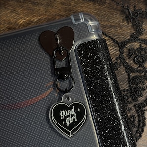 Op de afbeelding: Een doorzichtige telefoonhoes met een zwarte glitterrand en een zwart hartvormig bedeltje. De bedel heeft een zwart hartvormig hangertje met de woorden "good girl" in witte letters. De bedel is met een zwarte metalen sluiting aan de hoes bevestigd.