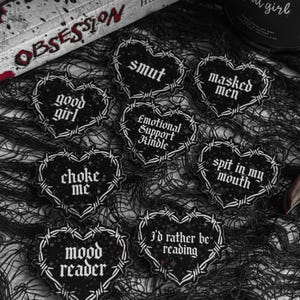 May include: A collection of black heart-shaped stickers with white text and barbed wire borders. The stickers feature phrases like "good girl," "smut," and "I'd rather be reading." The stickers are arranged on a dark, textured surface.