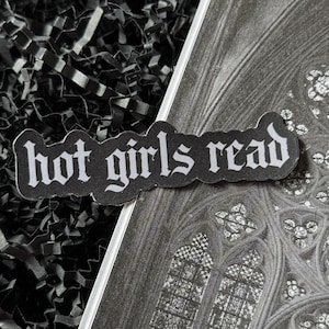 May include: A black sticker with the words "hot girls read" in white gothic font. The sticker is placed on a grayscale image of a stained-glass window and black shredded paper.