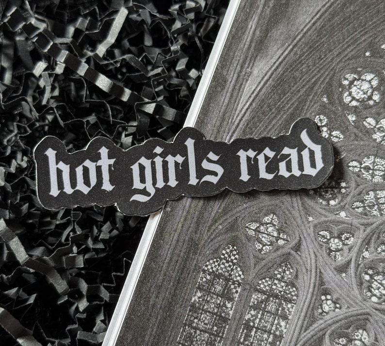 May include: A black sticker with the words "hot girls read" in white gothic font. The sticker is placed on a grayscale image of a stained-glass window and black shredded paper.