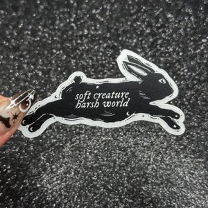 May include: A black and white sticker featuring a running rabbit silhouette. The sticker has the text "soft creature, harsh world" in a handwritten font. The sticker is on a glittery, dark gray background.