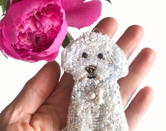 May include: A white beaded dog ornament with a pearl necklace. The ornament is held in a hand against a white background.