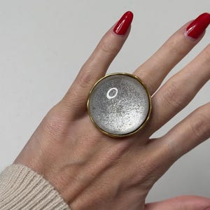 May include: A ring featuring a large, circular, convex centrepiece with a textured, metallic silver finish. The centrepiece is encased in a smooth, polished gold-tone metal rim that creates a distinct border around the reflective, shimmering surface.
