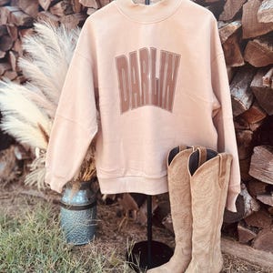May include: A light peach-colored crewneck sweatshirt featuring the word DARLIN printed in a bold, dark brown arched font across the chest, paired with a pair of tan suede knee-high boots with intricate decorative stitching along the shaft.