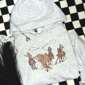 May include: A grey hooded sweatshirt with a graphic print of cowboys roping a brown bull. The cowboys are wearing brown hats and riding brown horses. The bull is brown with white markings.
