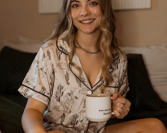 Puede incluir: Una mujer con una camiseta de pijama blanca y marrón con estampado de cactus se sienta en una cama sosteniendo una taza blanca que dice "You are my favorite".