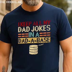 May include: Navy blue t-shirt with the text "I KEEP ALL MY DAD JOKES IN A DAD-A-BASE" in a retro font. The design includes a graphic of a database server. The shirt is a classic crew neck.