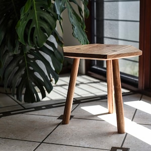 May include: A hexagonal wooden stool with three legs. The stool is made of light brown wood and has a natural finish. The stool is sitting on a tiled floor.