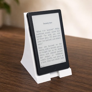 May include: A white plastic e-reader stand holding a black e-reader device. The screen displays text starting with the word Twenty-two and includes the sentence What I've learned, Molly muttered, mostly under her breath. The stand has a slanted back.