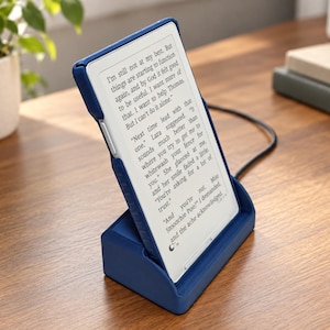 May include: A blue e-reader device sits in a matching blue charging dock. The screen displays black text on a white background, featuring dialogue from a book. A black charging cable extends from the back of the dock across the surface.