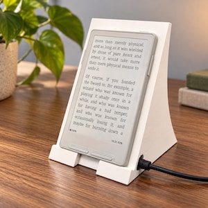 May include: A white angled e-reader stand holding a light gray e-reader device. The screen displays text about a sword and a wizard. A black charging cable is plugged into the side of the device. The stand has a solid, triangular profile.